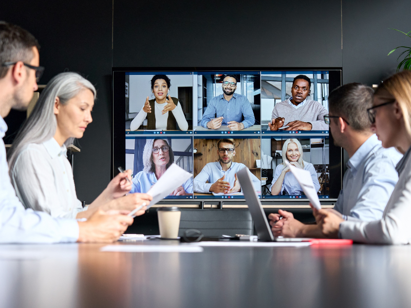 Group of employees sitting at a conference table with a virtual conference screen in the background